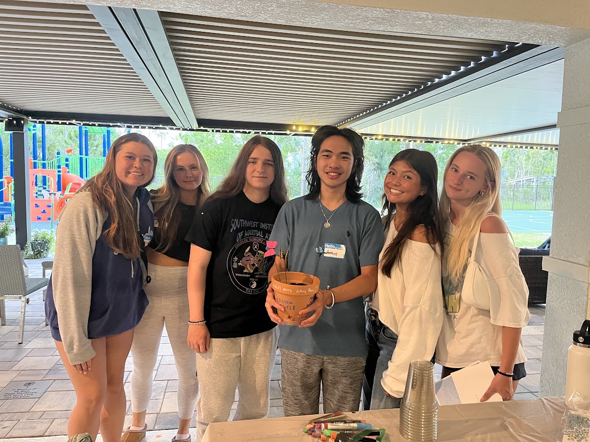 Group of teen volunteers smiling together at Valerie’s House while hosting an activity table with craft supplies during a group night event